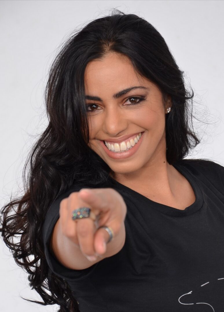 mulher morena, sorrindode camisa preta apontando o dedo cabelos longos e ondulados castanho escuro olhos castanhos brinco de stras