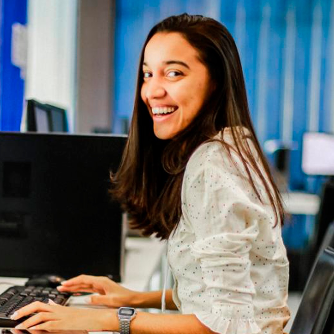 mulher morena clara cabelos castanhos medios longos sentada diante do computador com as maos no teclado camisa branca sorrindo
