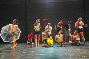 jovens com figurinos fazendo apresentaçao de dança com objetos guarda chuva, bacia e balao de coraçao
