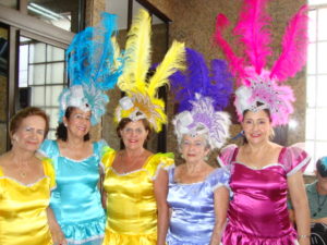 cinco mulheres com vestidos de fantasia e adereço de cabeça com plumas amarelo azul liás e rosa pink
