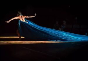 figurino de espetáculo mulher de costas musculosas braços abertos com um vestido azul com uma calda enorme caminhando