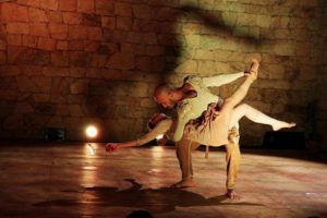 figurino de espetáculo homem negro com mulher branca no colo dançando no palco vestidos com figurino cor terrosos
