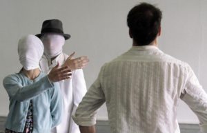 figurino de espetáculo mulher com rosto tampado com tecido branco homem com o rosto tampado com tecido branco apontando para um homem de costa caminhando com camisa branca