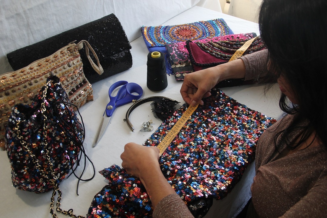 mulher fazendo bolsas artesanais preparando a modelagem de uma bolsa com um tecido de lantejoulas e várias bolsas prontas sobre a mesa